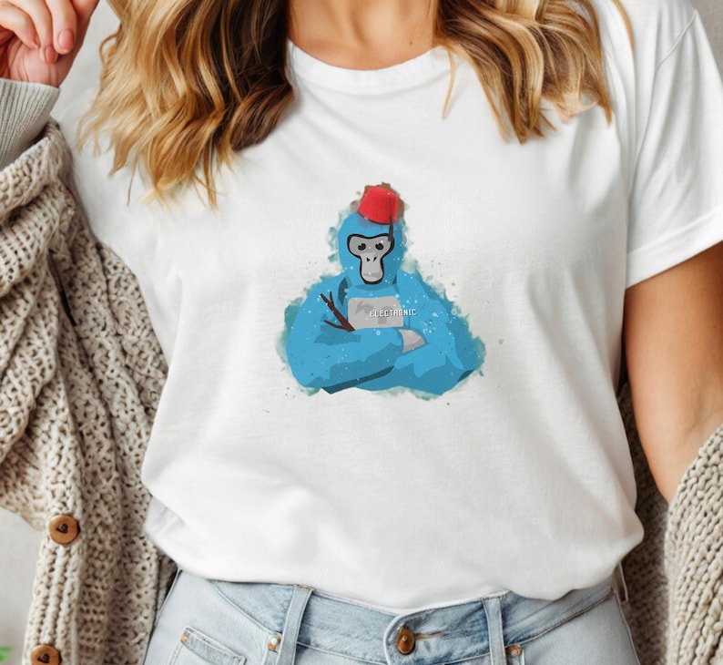 May include: White t-shirt featuring a blue watercolor illustration of a monkey wearing a red fez and holding a stick. The monkey has its arms crossed and the word "electronic" is printed on a small sign.