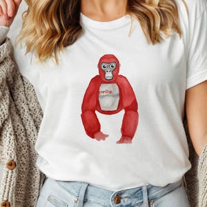 May include: White t-shirt featuring a red watercolor gorilla illustration. The gorilla has a white belly and the word "gorilla" printed on it. The shirt is made of a soft material.