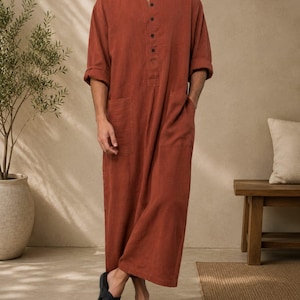 Op de afbeelding: Een lang, tot op de grond reikend gewaad met lange mouwen in een effen terracotta kleur. Het kledingstuk is voorzien van een mandarijnkraag, een gedeeltelijke knoopsluiting met vijf donkere knopen en twee grote opgestikte zakken aan de voorzijde. De mouwen zijn opgerold.
