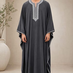 Könnte beinhalten: Ein dunkelgrauer Kaftan mit einer langen, fließenden Silhouette, weiten Ärmeln und einem silberfarben bestickten Ausschnitt. Das Kleidungsstück ist mit silbernen Paspeln an den Ärmelkanten und am Saum versehen und wird mit dunkelbraunen Sandalen kombiniert.