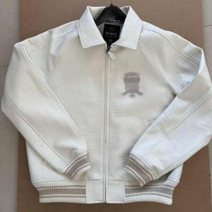 K&ouml;nnte beinhalten: Wei&szlig;e Lederjacke mit strukturierter Krokodilmusterung. Sie hat einen Rei&szlig;verschluss vorne, gerippte B&uuml;ndchen und Saum mit grauen Streifen und ein kleines Emblem auf der Brust. Die Jacke h&auml;ngt an einem Metallb&uuml;gel.