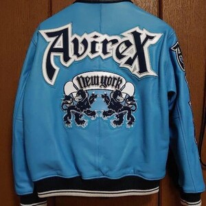 Peut inclure: Veste en cuir bleu clair avec le nom de la marque "Avirex" en lettres blanches et bleu marine. Le dos de la veste affiche le texte "New York" dans une banni&egrave;re blanche avec deux graphiques de lions noirs. La veste a des bordures noires et blanches.