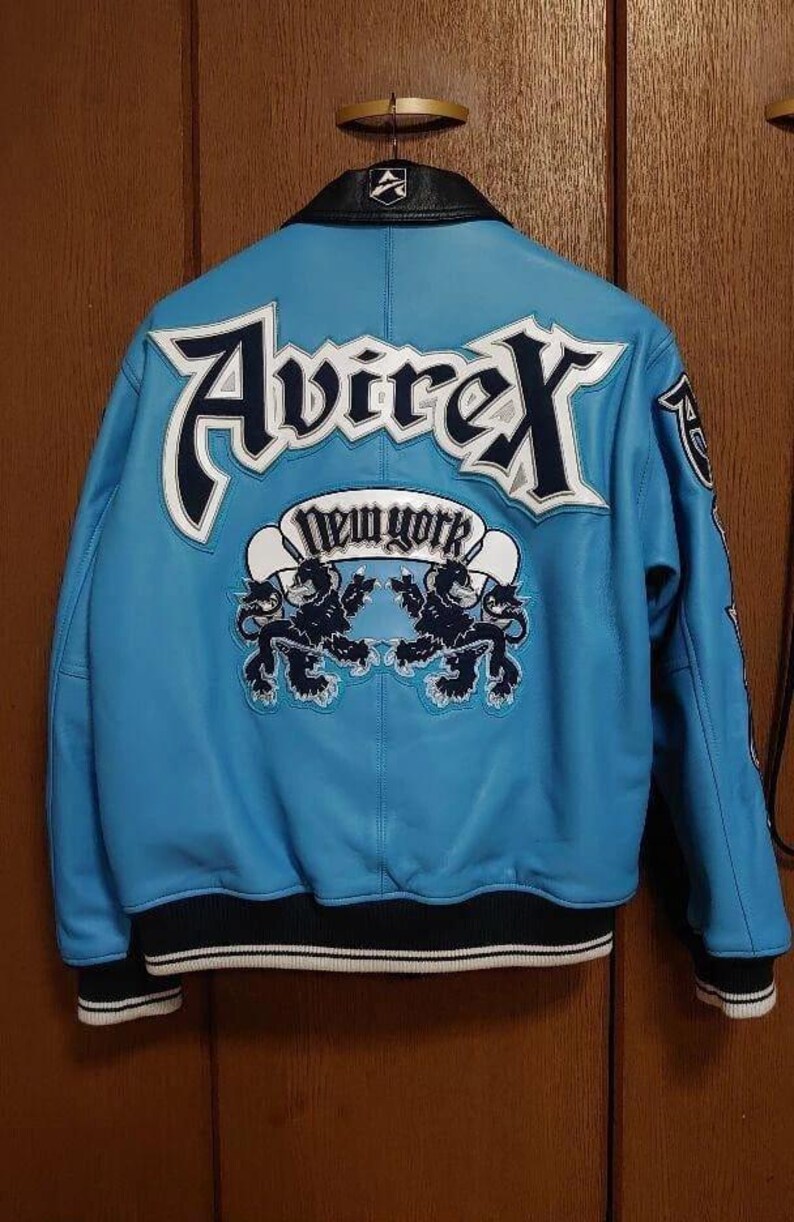 Peut inclure: Veste en cuir bleu clair avec le nom de la marque "Avirex" en lettres blanches et bleu marine. Le dos de la veste affiche le texte "New York" dans une banni&egrave;re blanche avec deux graphiques de lions noirs. La veste a des bordures noires et blanches.