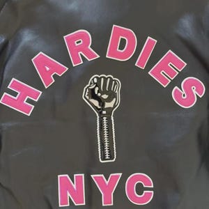 Puede incluir: Chaqueta negra con la palabra "HARDIES" en letras rosas con contornos blancos, arqueada en la parte superior. Debajo, un gr&aacute;fico de un pu&ntilde;o negro sosteniendo una cremallera. La palabra "NYC" est&aacute; en la parte inferior en letras rosas con contornos blancos.