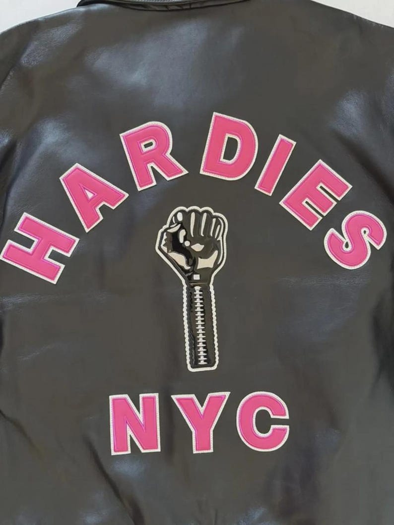 Puede incluir: Chaqueta negra con la palabra "HARDIES" en letras rosas con contornos blancos, arqueada en la parte superior. Debajo, un gr&aacute;fico de un pu&ntilde;o negro sosteniendo una cremallera. La palabra "NYC" est&aacute; en la parte inferior en letras rosas con contornos blancos.
