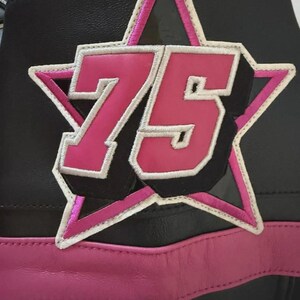 Puede incluir: Detalle de una chaqueta de cuero negra con un parche de estrella rosa con el n&uacute;mero 75. El parche tiene un contorno blanco y est&aacute; cosido a la chaqueta. Una franja de cuero rosa recorre la parte inferior.
