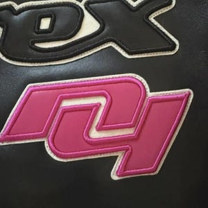 Puede incluir: Primer plano de una superficie negra con dos parches bordados. El parche superior es negro con ribete blanco y negro, y el parche inferior es rosa con ribete blanco y rosa. Los parches parecen ser letras.