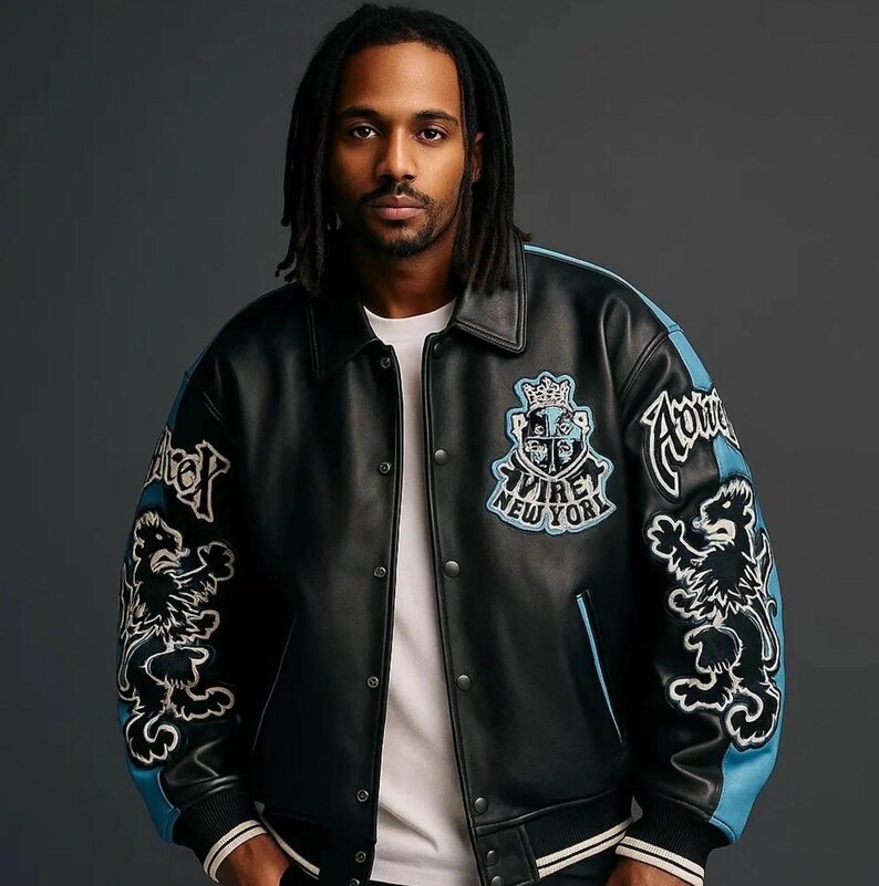 Peut inclure: Veste en cuir noire avec des d&eacute;tails et des &eacute;cussons bleus. La veste a une fermeture boutonn&eacute;e, un col et des &eacute;cussons avec du texte et un motif de lion. Le texte sur la veste dit "AVIRE NEW YORK".