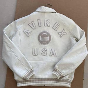 K&ouml;nnte beinhalten: Cremefarbene Lederjacke mit gepr&auml;gter Krokodiltextur. Auf dem R&uuml;cken steht das Wort "AVIREX" &uuml;ber einem kreisf&ouml;rmigen Emblem und darunter "USA". Gerippte B&uuml;ndchen und Saum in kontrastierendem Beige.