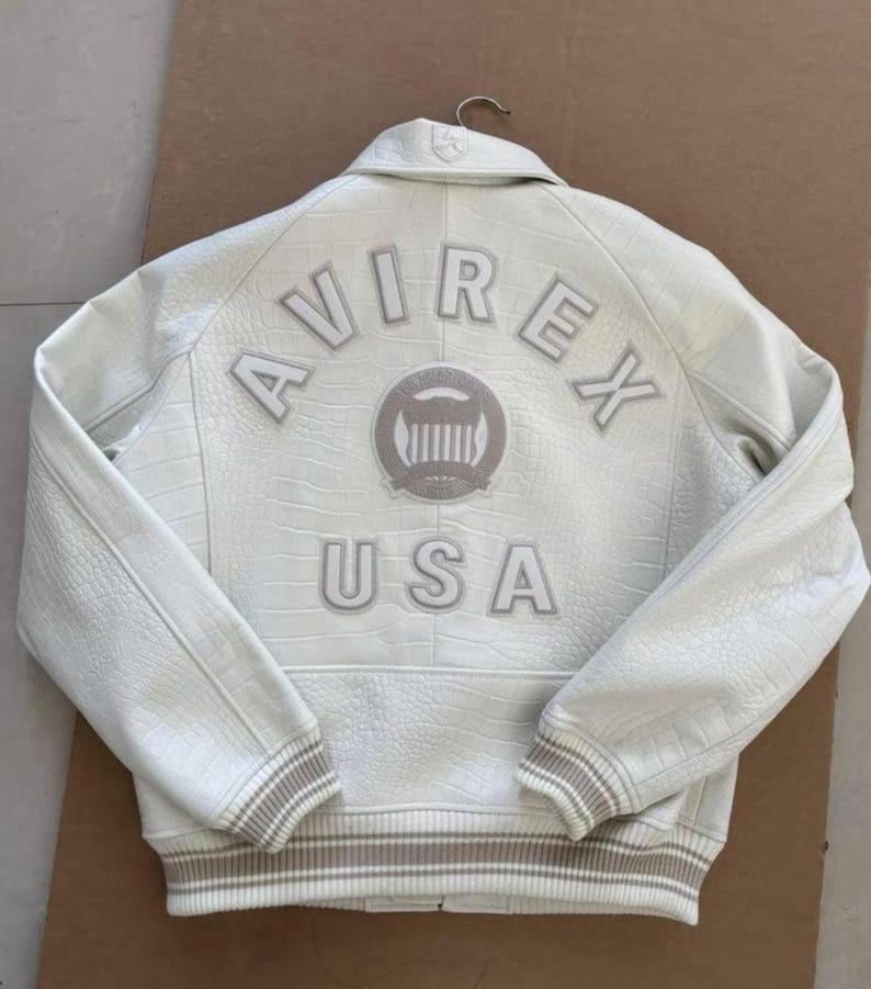 K&ouml;nnte beinhalten: Cremefarbene Lederjacke mit gepr&auml;gter Krokodiltextur. Auf dem R&uuml;cken steht das Wort "AVIREX" &uuml;ber einem kreisf&ouml;rmigen Emblem und darunter "USA". Gerippte B&uuml;ndchen und Saum in kontrastierendem Beige.