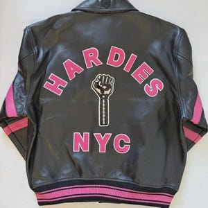 Puede incluir: Chaqueta de cuero negra con detalles rosas en las mangas y el bajo. La parte trasera presenta la palabra "HARDIES" en letras rosas sobre un pu&ntilde;o en blanco y negro, con "NYC" debajo.