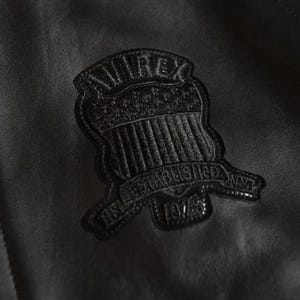 May include: Close-up of a black leather jacket with a textured surface. A black patch is sewn onto the jacket, featuring the word "AVIREX" above a shield design and the text "ESTABLISHED NAVY 1975".