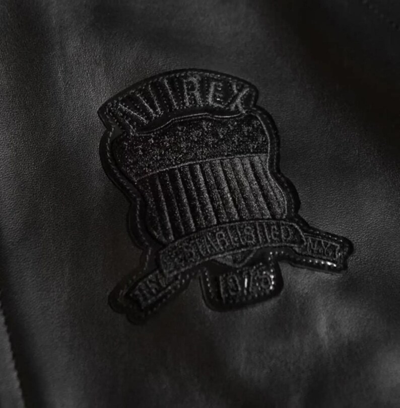 May include: Close-up of a black leather jacket with a textured surface. A black patch is sewn onto the jacket, featuring the word "AVIREX" above a shield design and the text "ESTABLISHED NAVY 1975".