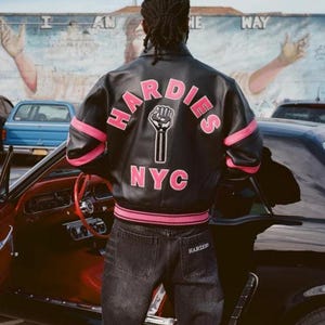 Puede incluir: Chaqueta de cuero negra con detalles rosas y el texto "HARDIES NYC" en la espalda. La chaqueta presenta un gr&aacute;fico de un pu&ntilde;o en alto. La persona lleva vaqueros oscuros con la palabra "HARDIES" en el bolsillo trasero.