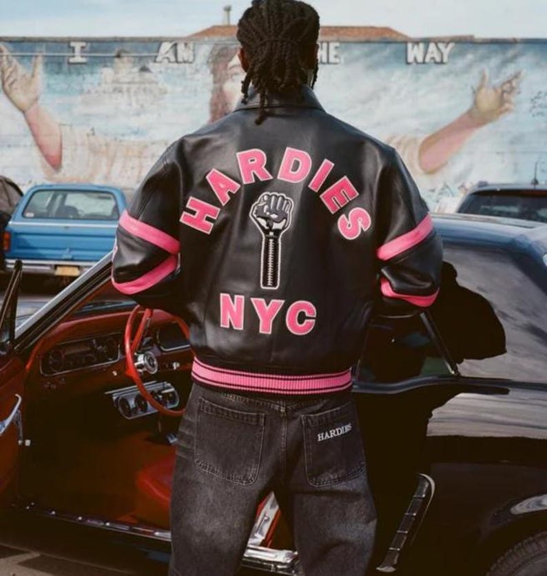 Puede incluir: Chaqueta de cuero negra con detalles rosas y el texto "HARDIES NYC" en la espalda. La chaqueta presenta un gr&aacute;fico de un pu&ntilde;o en alto. La persona lleva vaqueros oscuros con la palabra "HARDIES" en el bolsillo trasero.