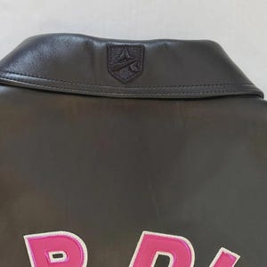 Puede incluir: Primer plano del cuello de una chaqueta de cuero gris oscuro con un peque&ntilde;o emblema. La chaqueta tiene un dise&ntilde;o rosa y blanco en la parte delantera. El cuello es de un tono gris m&aacute;s oscuro que el resto de la chaqueta.