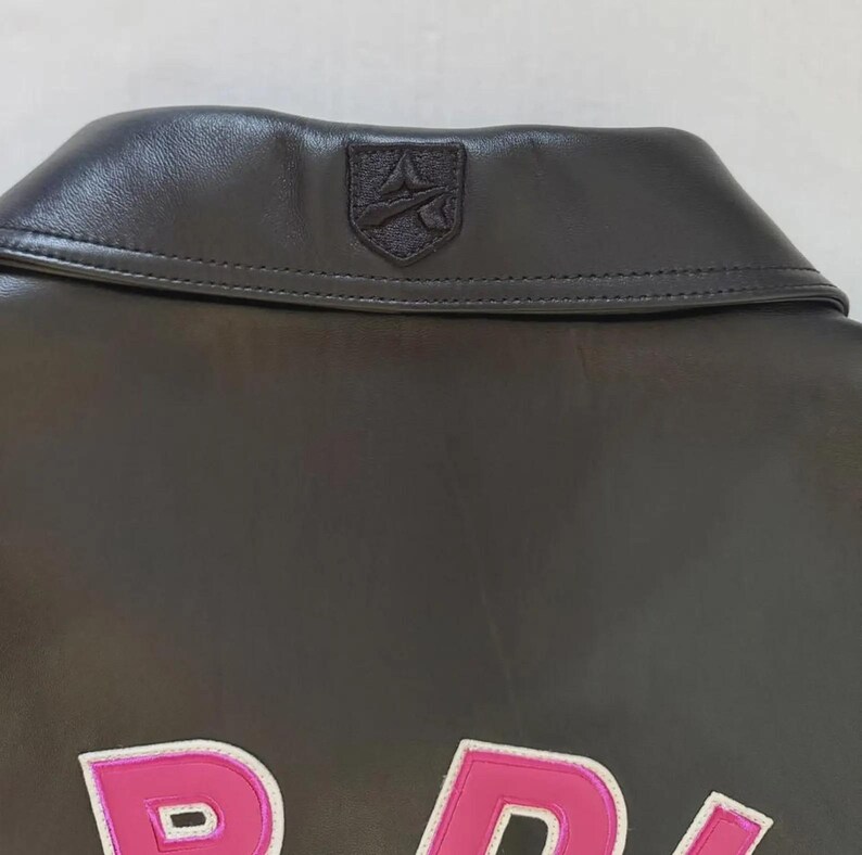 Puede incluir: Primer plano del cuello de una chaqueta de cuero gris oscuro con un peque&ntilde;o emblema. La chaqueta tiene un dise&ntilde;o rosa y blanco en la parte delantera. El cuello es de un tono gris m&aacute;s oscuro que el resto de la chaqueta.
