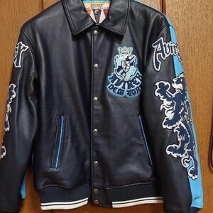 Peut inclure: Veste en cuir bleu marine avec des d&eacute;tails bleu clair et blancs. La veste pr&eacute;sente un &eacute;cusson et des lettres sur la poitrine et les manches. Le col et les poignets sont c&ocirc;tel&eacute;s avec des rayures blanches. La veste est suspendue &agrave; un cintre noir.