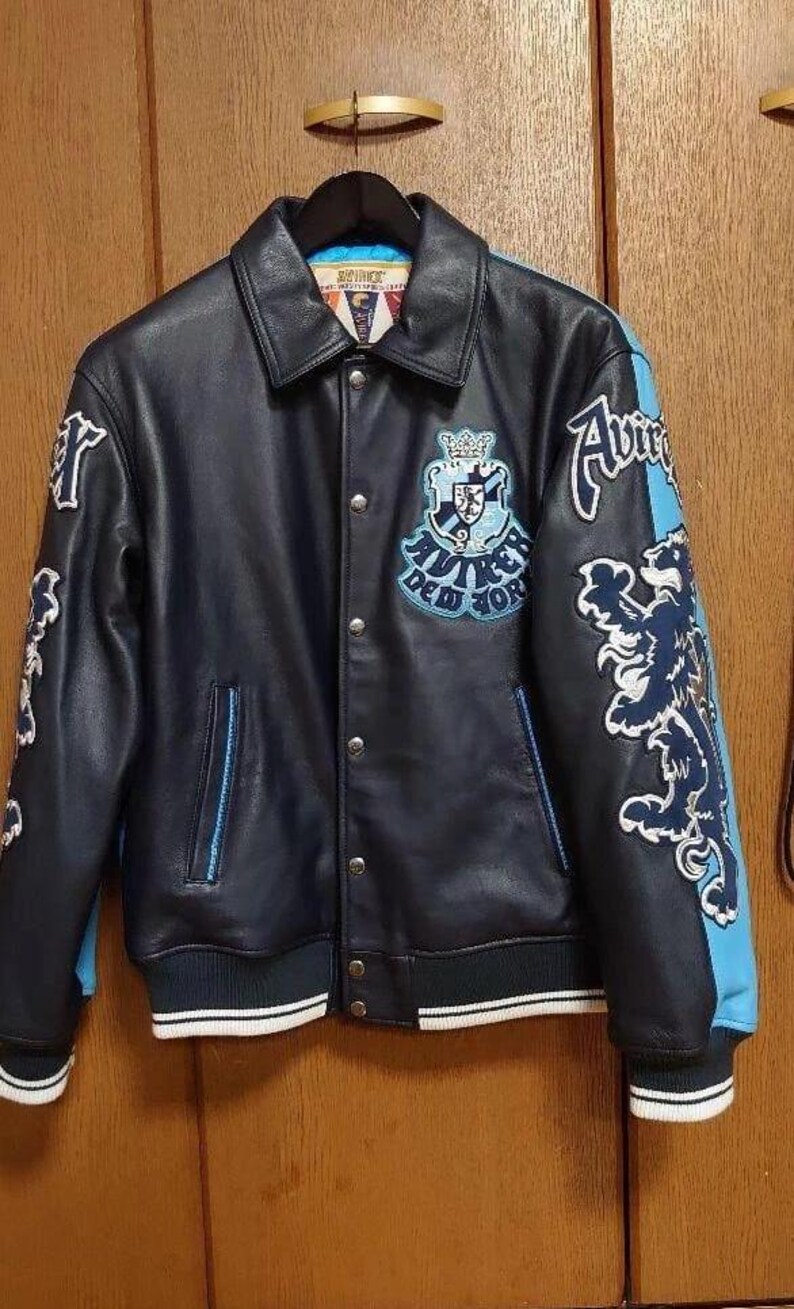 Peut inclure: Veste en cuir bleu marine avec des d&eacute;tails bleu clair et blancs. La veste pr&eacute;sente un &eacute;cusson et des lettres sur la poitrine et les manches. Le col et les poignets sont c&ocirc;tel&eacute;s avec des rayures blanches. La veste est suspendue &agrave; un cintre noir.