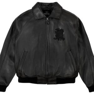 May include: Black leather bomber jacket with a zip-up front, collar, and two front pockets. The jacket features a black emblem on the chest and black ribbed cuffs and hem. The jacket is a classic style.