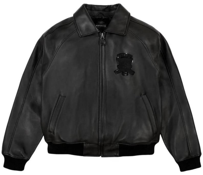 May include: Black leather bomber jacket with a zip-up front, collar, and two front pockets. The jacket features a black emblem on the chest and black ribbed cuffs and hem. The jacket is a classic style.