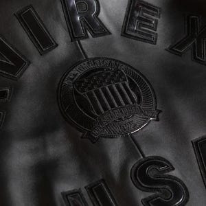 May include: Close-up of a black leather jacket with raised lettering and patches. The jacket features the word "AIREX" and a circular patch with "AMERICAN ESTABLISHED 1975" text and a flag design. The jacket is made of a smooth, shiny leather.
