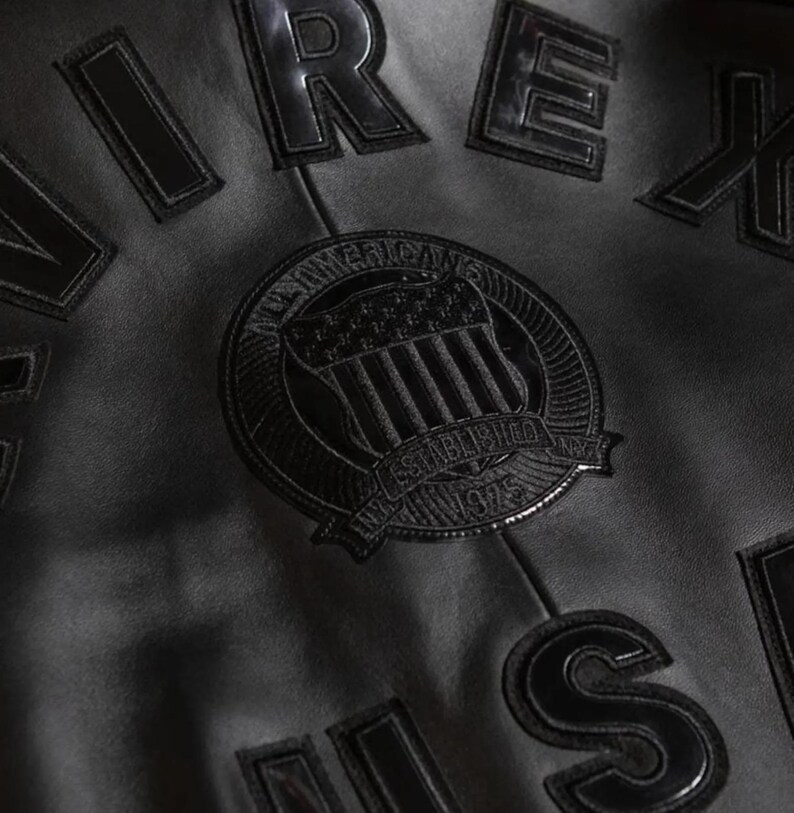 May include: Close-up of a black leather jacket with raised lettering and patches. The jacket features the word "AIREX" and a circular patch with "AMERICAN ESTABLISHED 1975" text and a flag design. The jacket is made of a smooth, shiny leather.