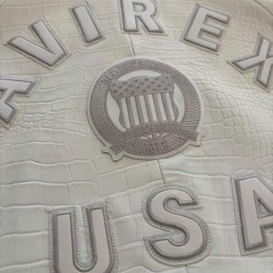 K&ouml;nnte beinhalten: Cremefarbene Lederjacke mit gepr&auml;gter Krokodiltextur. Mit den Worten "VIREX" und "USA" in erhabenen Buchstaben. Ein kreisf&ouml;rmiger Aufn&auml;her mit einem amerikanischen Flaggen-Design ist ebenfalls sichtbar. Die Jacke hat eine klassische, Vintage-&Auml;sthetik.