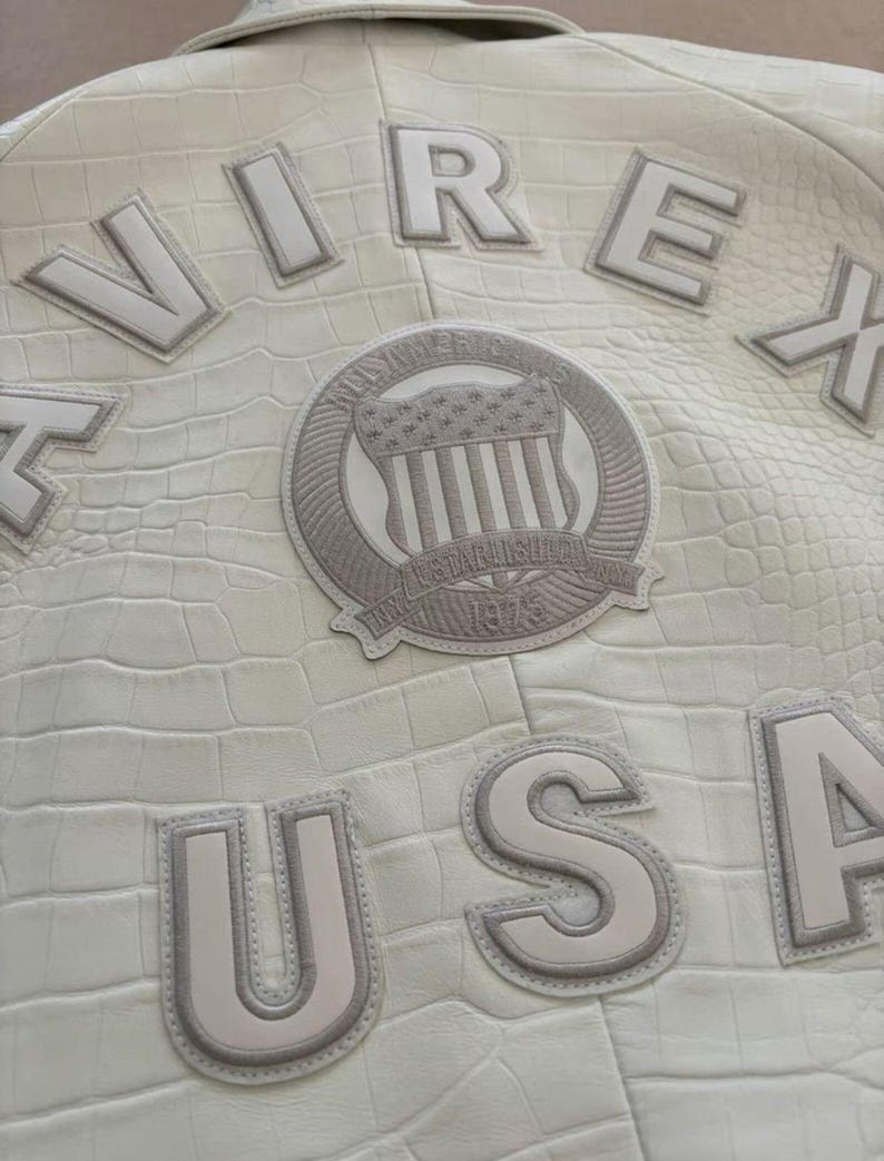 K&ouml;nnte beinhalten: Cremefarbene Lederjacke mit gepr&auml;gter Krokodiltextur. Mit den Worten "VIREX" und "USA" in erhabenen Buchstaben. Ein kreisf&ouml;rmiger Aufn&auml;her mit einem amerikanischen Flaggen-Design ist ebenfalls sichtbar. Die Jacke hat eine klassische, Vintage-&Auml;sthetik.