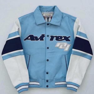 Op de afbeelding: Een lichtblauwe Avi'rex varsity jack met witte mouwen en marineblauwe accenten. De jas heeft het Avi'rex-logo in marineblauw en wit, met een knoopsluiting en een gestreepte zoom en manchetten.