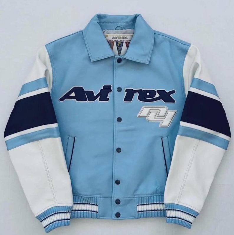 Op de afbeelding: Een lichtblauwe Avi'rex varsity jack met witte mouwen en marineblauwe accenten. De jas heeft het Avi'rex-logo in marineblauw en wit, met een knoopsluiting en een gestreepte zoom en manchetten.