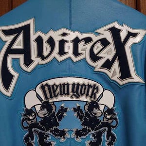 Peut inclure: Veste en cuir bleu clair avec le mot "Avirex" en grandes lettres blanches et bleu marine dans le dos. En dessous, une banni&egrave;re blanche avec les mots "New York" et deux embl&egrave;mes de lions noirs.