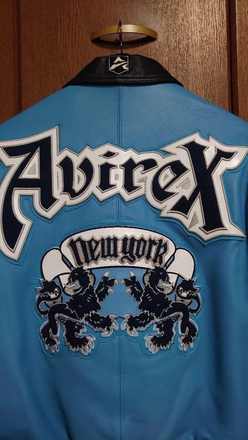 Peut inclure: Veste en cuir bleu clair avec le mot "Avirex" en grandes lettres blanches et bleu marine dans le dos. En dessous, une banni&egrave;re blanche avec les mots "New York" et deux embl&egrave;mes de lions noirs.
