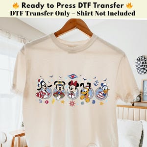 May include: A cream-coloured t-shirt with a nautical-themed graphic of cartoon characters. The graphic includes images of Goofy, Mickey Mouse, Minnie Mouse, Pluto, and Donald Duck in circular frames with nautical elements. Text at the top reads "Ready to Press DTF Transfer".