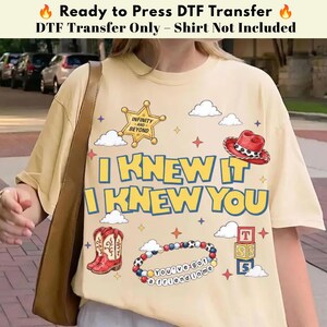 May include: A cream-colored t-shirt featuring a graphic print with the text I KNEW IT I KNEW YOU, a gold sheriff badge with INFINITY AND BEYOND, a red cowboy hat, red cowboy boots, a beaded bracelet with YOU'VE GOT A FRIEND IN ME, and blocks.