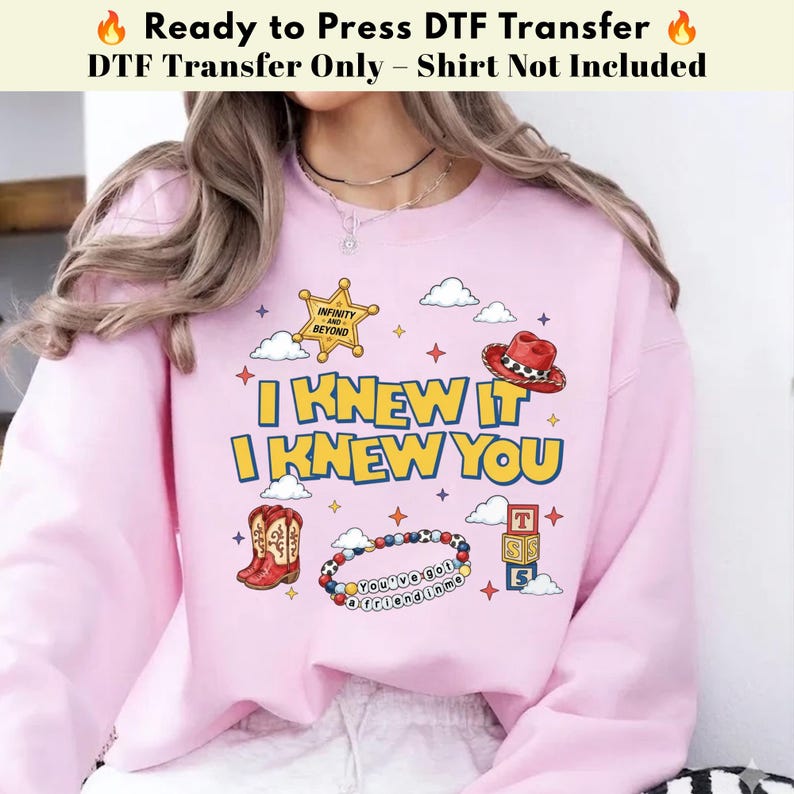 May include: A light pink sweatshirt featuring a graphic print with the text I KNEW IT I KNEW YOU. The design includes a yellow sheriff star, a red cowboy hat, brown cowboy boots, a beaded bracelet with text, and stacked alphabet blocks.