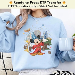 Pode incluir: Camisola azul clara com uma transfer&ecirc;ncia DTF do Mickey Mouse com uma roupa de feiticeiro, segurando uma varinha. O design inclui um castelo, um globo e o texto "Ready to Press DTF Transfer".