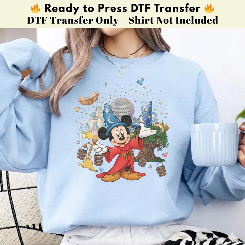 Pode incluir: Camisola azul clara com uma transfer&ecirc;ncia DTF do Mickey Mouse com uma roupa de feiticeiro, segurando uma varinha. O design inclui um castelo, um globo e o texto "Ready to Press DTF Transfer".