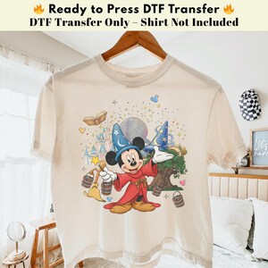 Pode incluir: Camiseta bege com um gr&aacute;fico colorido do Mickey Mouse com uma roupa de feiticeiro, segurando uma varinha m&aacute;gica. O design inclui um castelo, um globo e estrelas. O texto na parte superior diz "Ready to Press DTF Transfer" e "DTF Transfer Only - Shirt Not Included."