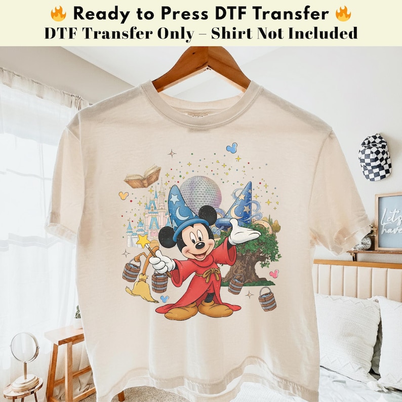 Pode incluir: Camiseta bege com um gr&aacute;fico colorido do Mickey Mouse com uma roupa de feiticeiro, segurando uma varinha m&aacute;gica. O design inclui um castelo, um globo e estrelas. O texto na parte superior diz "Ready to Press DTF Transfer" e "DTF Transfer Only - Shirt Not Included."