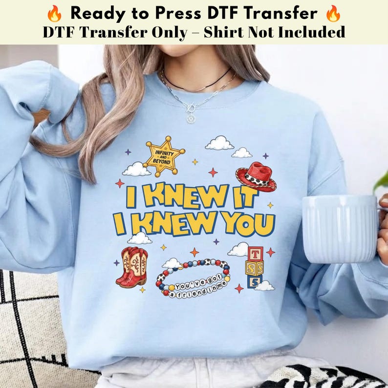 May include: A light blue sweatshirt featuring a graphic print with the text I KNEW IT I KNEW YOU, a yellow sheriff star with INFINITY AND BEYOND, a red cowboy hat, red cowboy boots, a beaded bracelet with YOU'VE GOT A FRIEND IN ME, and blocks.
