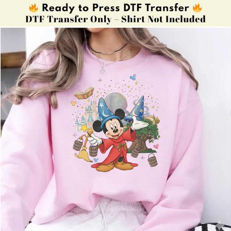Pode incluir: Um moletom rosa claro com um gr&aacute;fico colorido do Mickey Mouse em traje de feiticeiro, com um castelo, uma &aacute;rvore e o texto "Ready to Press DTF Transfer". O design inclui estrelas e elementos m&aacute;gicos.