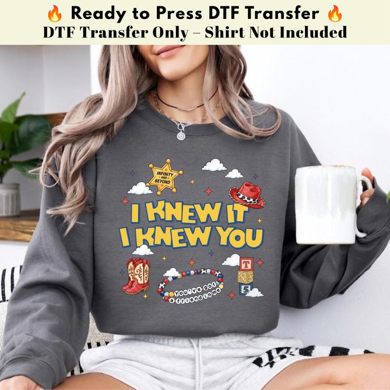 May include: A dark gray sweatshirt featuring a graphic print with the text I KNEW IT I KNEW YOU. The design includes a yellow sheriff star, a red cowboy hat, red cowboy boots, a beaded bracelet with text, and stacked alphabet blocks.
