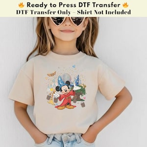 Pode incluir: Uma camiseta bege com um gr&aacute;fico colorido do Mickey Mouse vestido de mago. O gr&aacute;fico inclui um castelo, uma &aacute;rvore e um globo. O texto na parte superior diz "Ready to Press DTF Transfer".