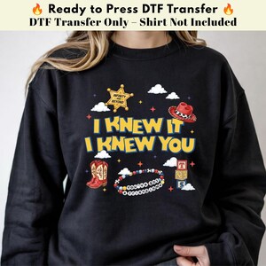May include: A black sweatshirt featuring a graphic print with the text I KNEW IT I KNEW YOU. The design includes a gold sheriff badge with the text INFINITY AND BEYOND, a red cowboy hat, brown cowboy boots, a beaded bracelet, and alphabet blocks.