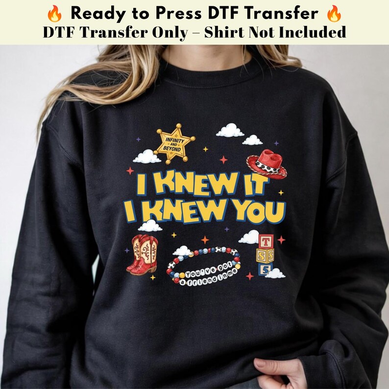 May include: A black sweatshirt featuring a graphic print with the text I KNEW IT I KNEW YOU. The design includes a gold sheriff badge with the text INFINITY AND BEYOND, a red cowboy hat, brown cowboy boots, a beaded bracelet, and alphabet blocks.