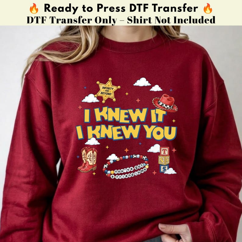 May include: A maroon sweatshirt featuring a graphic print with the text I KNEW IT I KNEW YOU, a yellow sheriff star badge labeled INFINITY AND BEYOND, a red cowboy hat, brown cowboy boots, a beaded bracelet, and a stack of alphabet blocks.