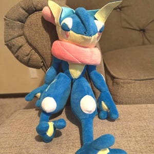 May include: A plush toy of a blue and yellow creature with large ears, a pink scarf, and white spots on its legs. The toy is sitting on a gray upholstered surface.