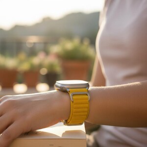 May include: A silver smartwatch with a bright yellow band is worn on a wrist. The band is made of a textured material. The watch is in focus, with a blurred background of plants and sunlight.