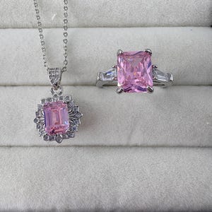 Könnte beinhalten: Eine silberfarbene Halskette mit Anhänger und ein passender Ring. Der Anhänger und der Ring zeigen beide einen großen, rechteckigen rosa Edelstein, der von kleineren, klaren Steinen umgeben ist. Der Ring hat außerdem klare Steine auf beiden Seiten des rosa Edelsteins.